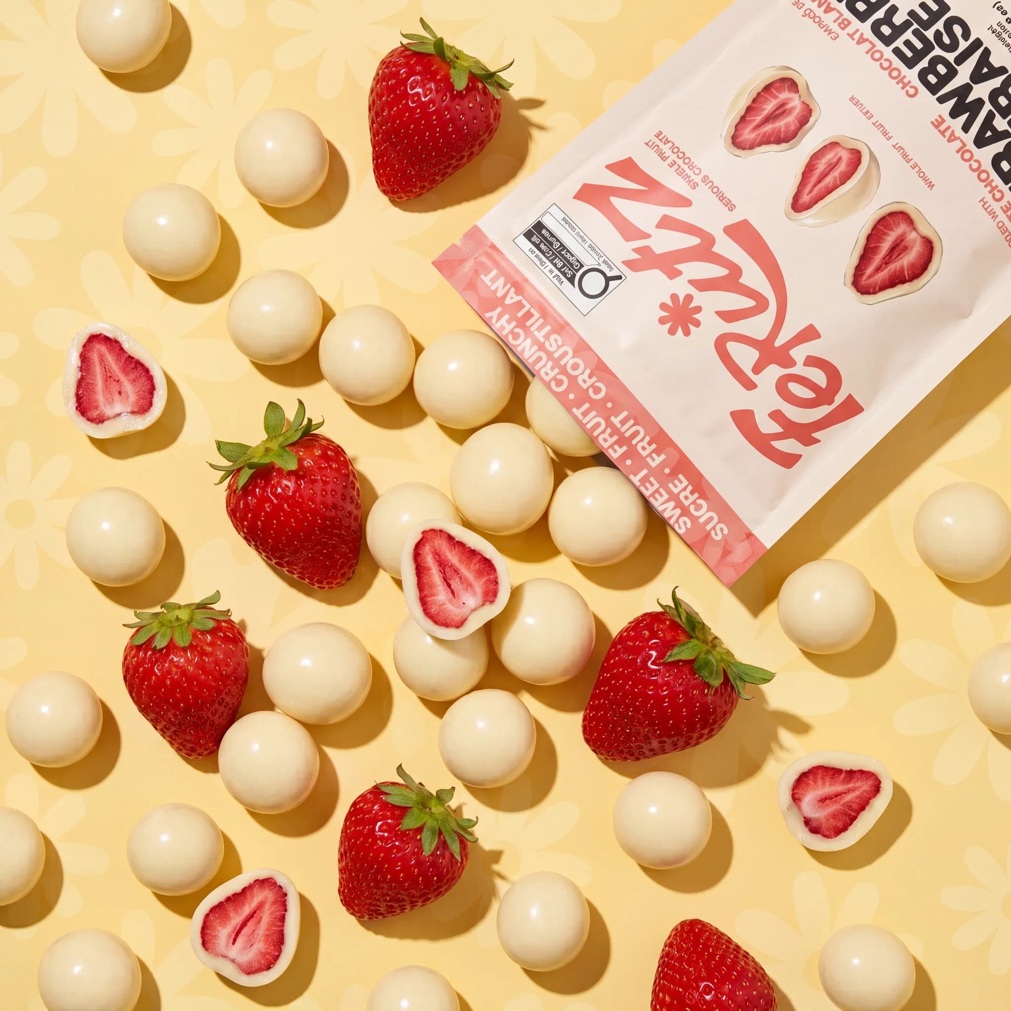 Strawberries and white chocolate bites scattered on butter yellow, the Ferutz bag tilting into frame